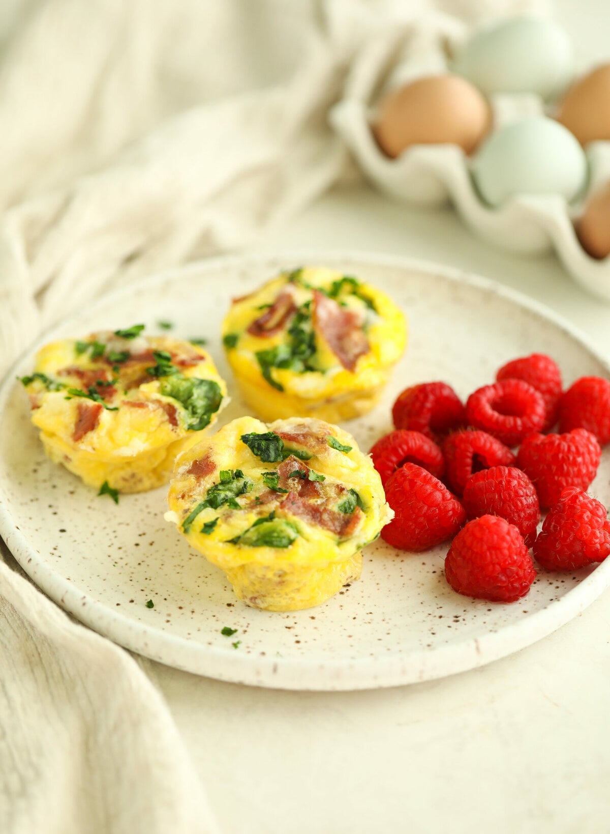 side view of 3 turkey bacon egg bites on a ceramic plate with a side of fresh raspberries - there are fresh eggs in a container in the background