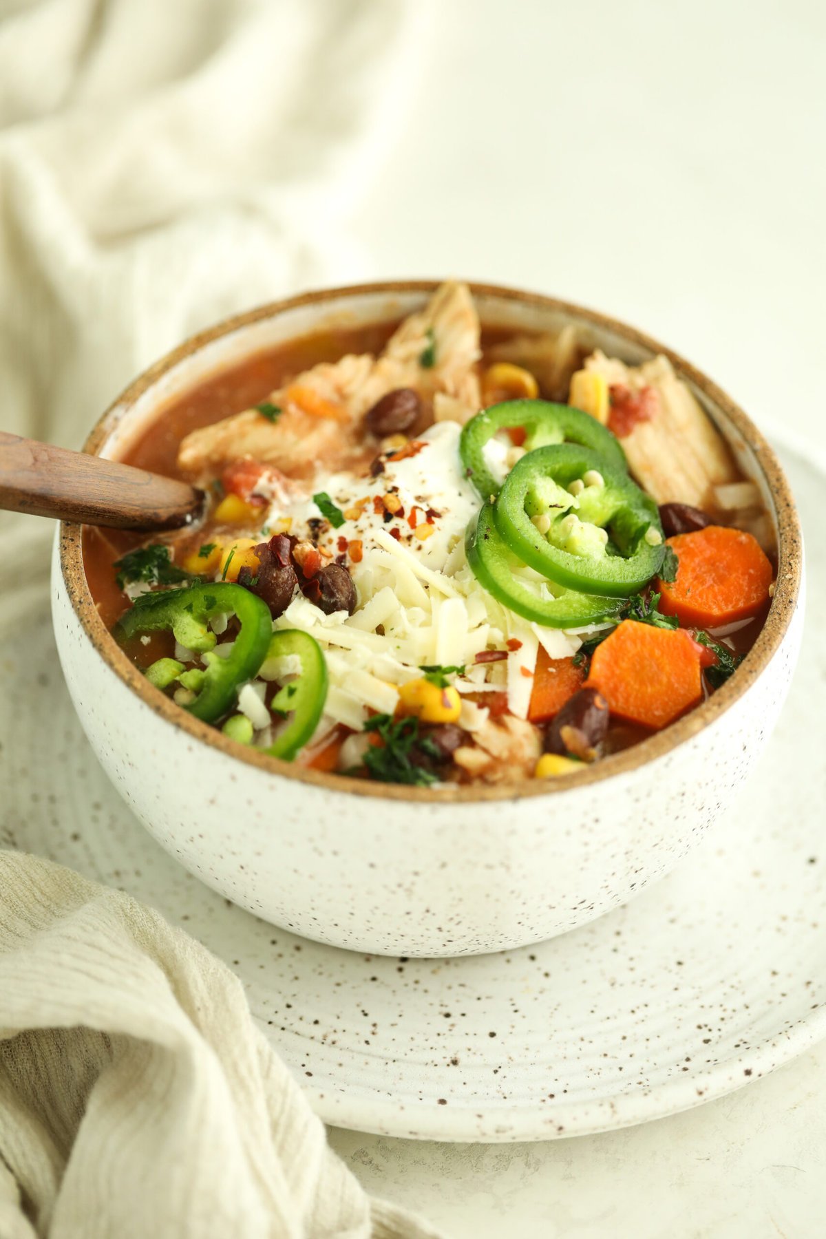 side view of a ceramic bowl sitting on a plate. The bowl is filled with chicken taco soup and topped with sour cream, jalapeños and cheese