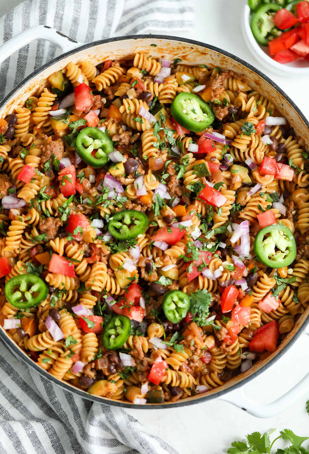 one pot taco pasta in a large ceramic skillet. It is topped with fresh cilantro, sliced jalapeños and diced tomatoes.