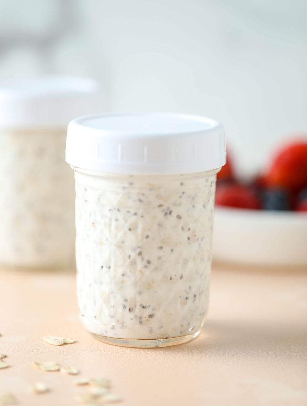 easy overnight oats in a glass jar with a white lid, there is a bowl of fresh berries behind it